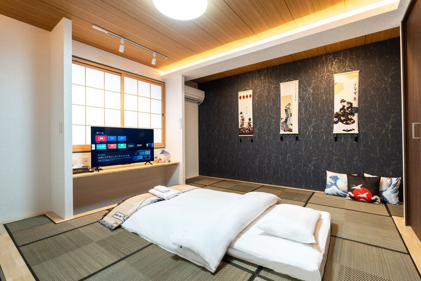 Japanese-style bedroom with tatami flooring, a futon bed, a TV, and decorative hanging scrolls on a dark wall.