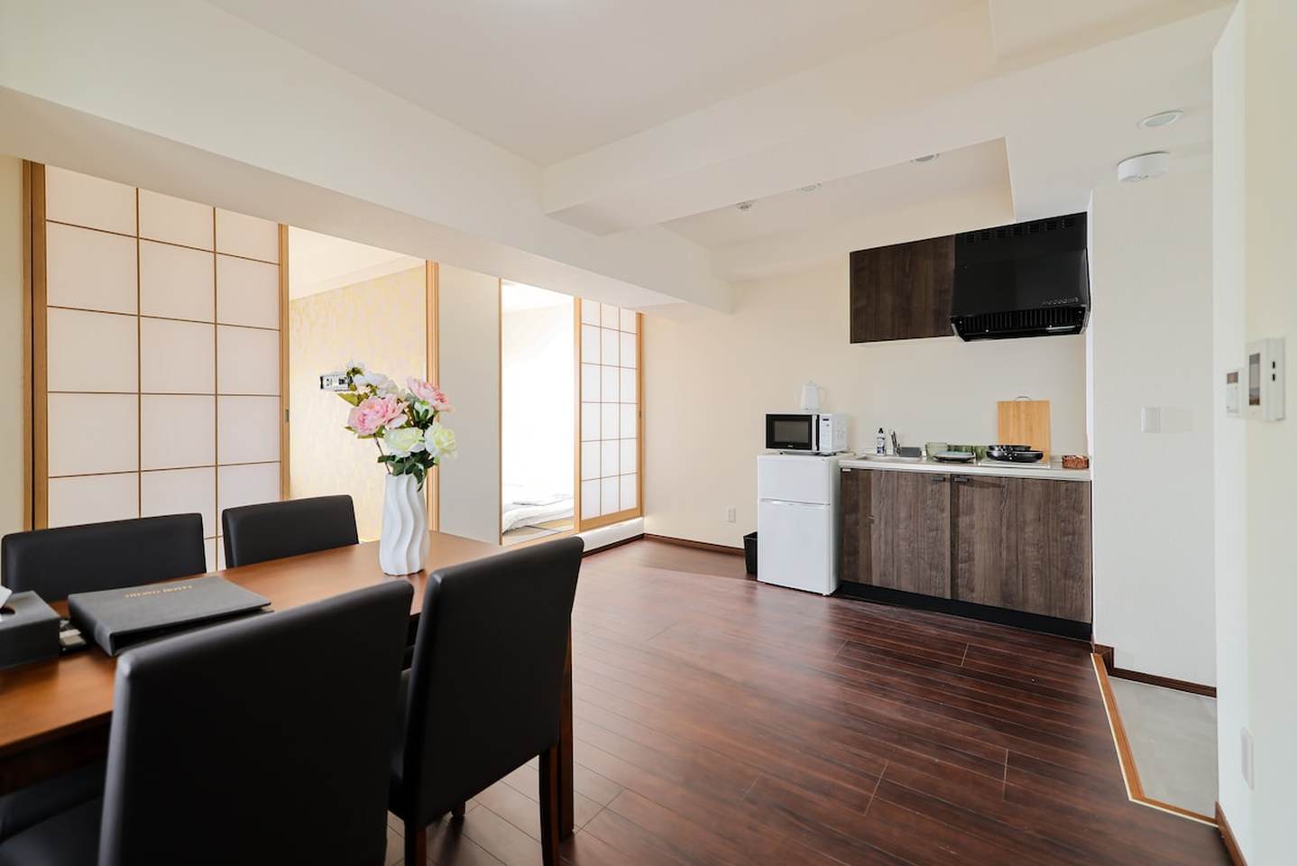 Dining table with black chairs next to a compact kitchen with dark wood cabinets and a mini-fridge.