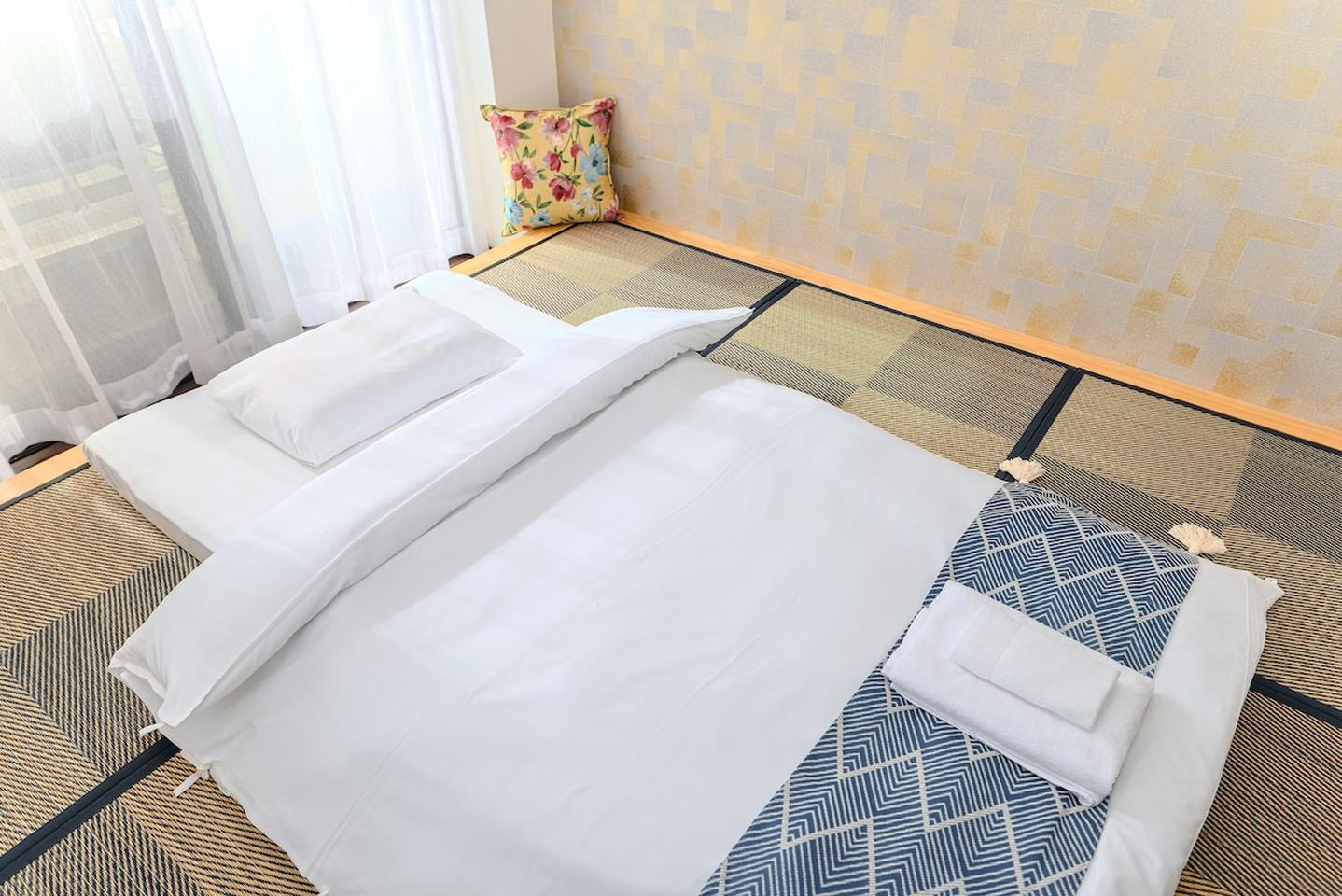 Traditional Japanese futon bed laid out on tatami mats with white linens and a patterned throw.