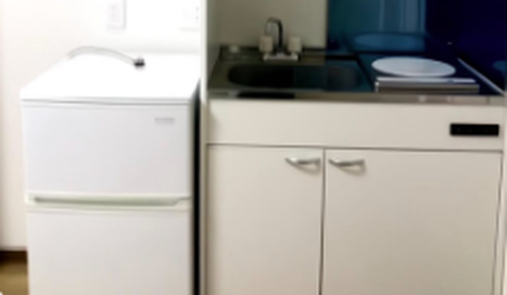 Compact kitchenette featuring a small white refrigerator, sink, and stovetop with under-counter cabinets.
