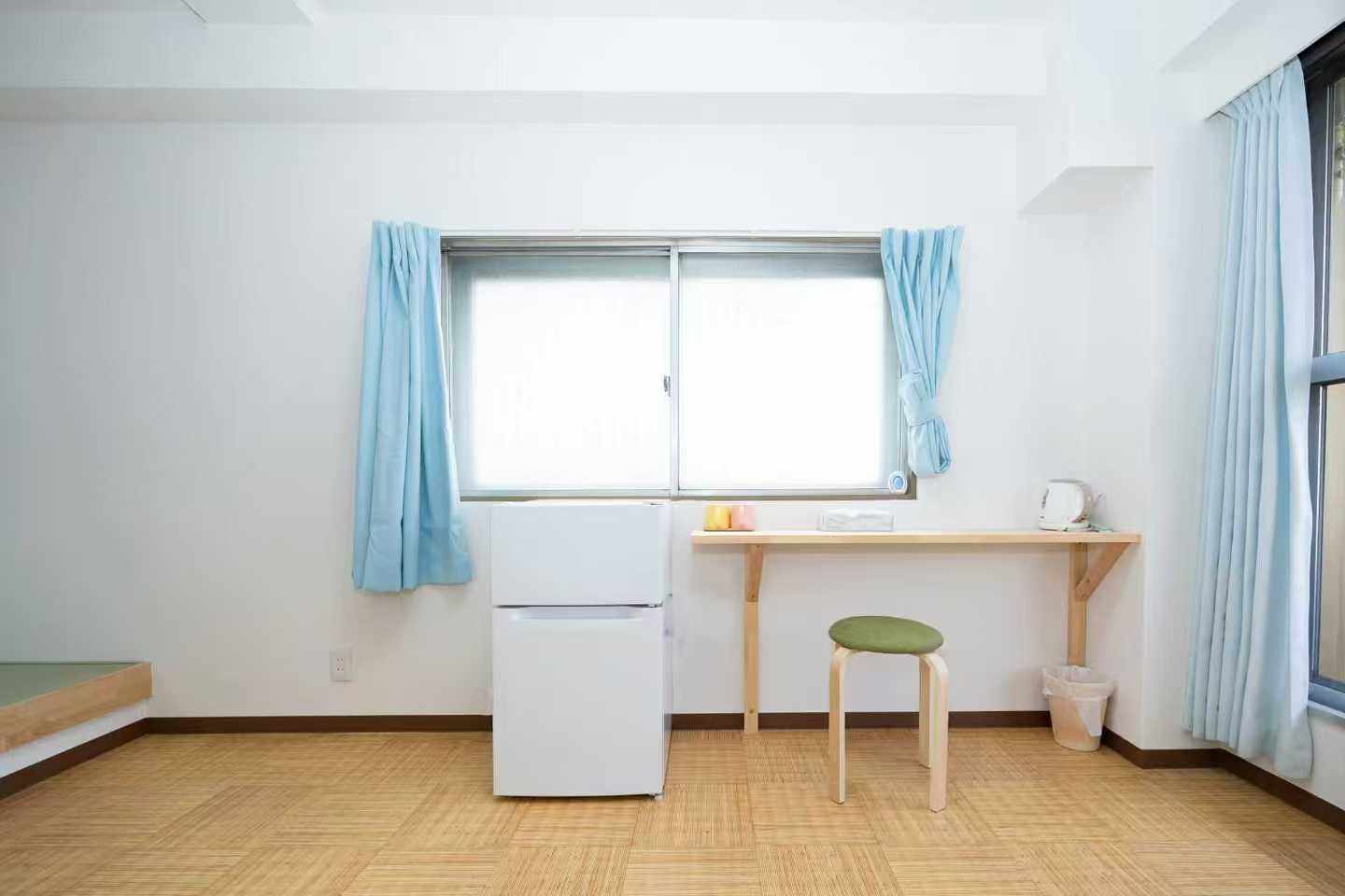 A small white refrigerator next to a wooden desk with a green stool under a window with light blue curtains.