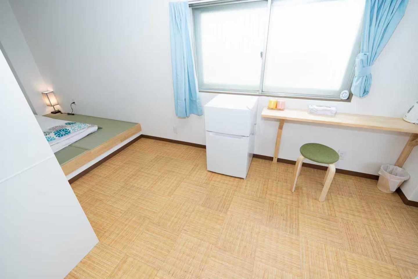 Compact room with a raised tatami sleeping platform, mini fridge, wooden desk, and green stool.