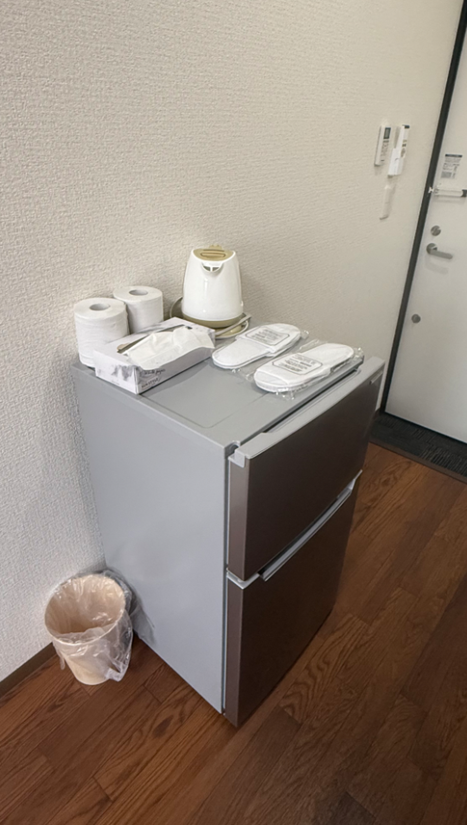 Small refrigerator with an electric kettle, tissues, toilet paper, and packaged slippers on top.