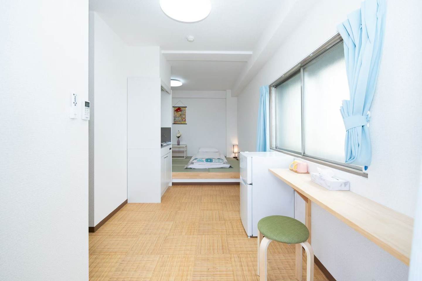 Narrow studio apartment with a wooden counter, green stool, small refrigerator, and a futon on a raised tatami platform.
