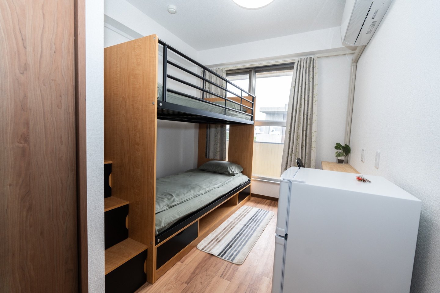 Compact bedroom featuring a wooden bunk bed with green bedding, a small white refrigerator, and a window.