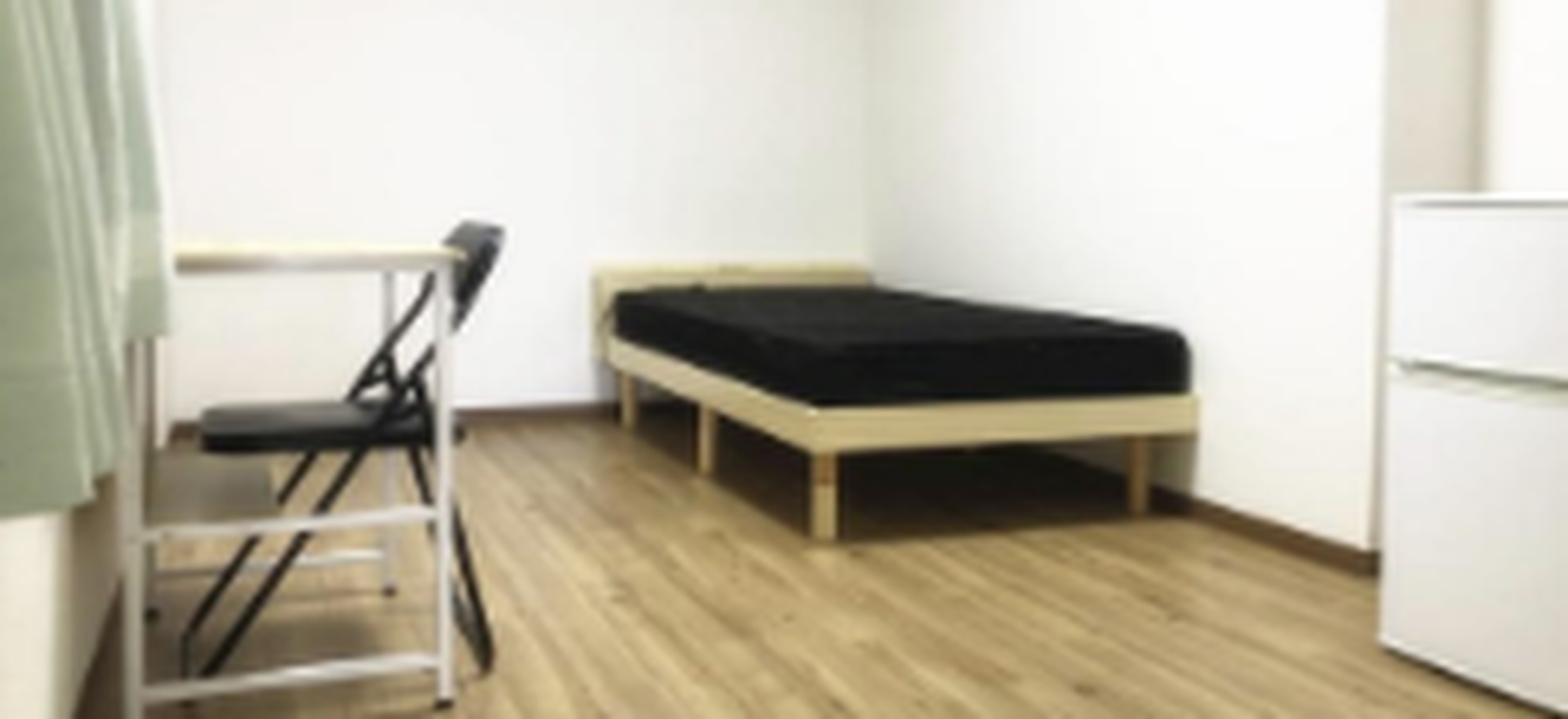 Minimalist bedroom with a single bed, a small desk and chair, and a mini fridge on wood flooring.