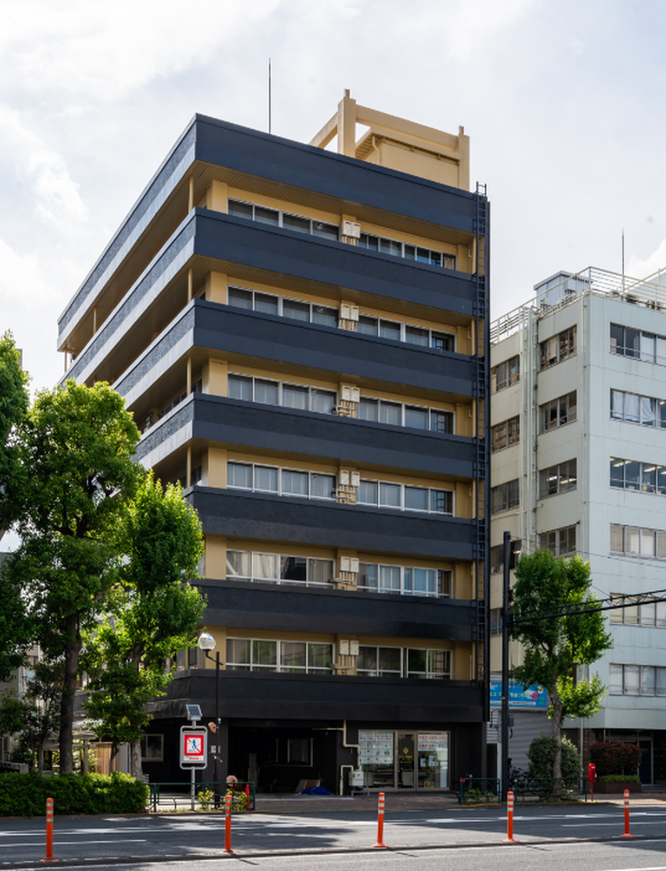 Exterior view of a multi-story black and beige building on a Tokyo street with green trees.