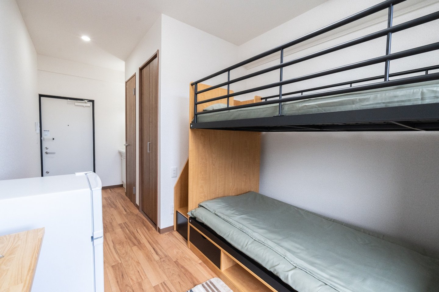 Compact studio apartment featuring a wooden bunk bed with green bedding and a small refrigerator.