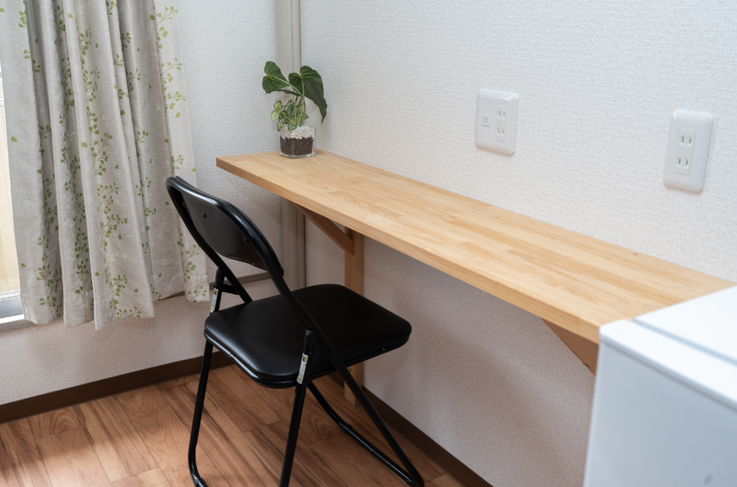 A narrow wooden wall-mounted desk with a black folding chair, a small potted plant, and wall outlets.