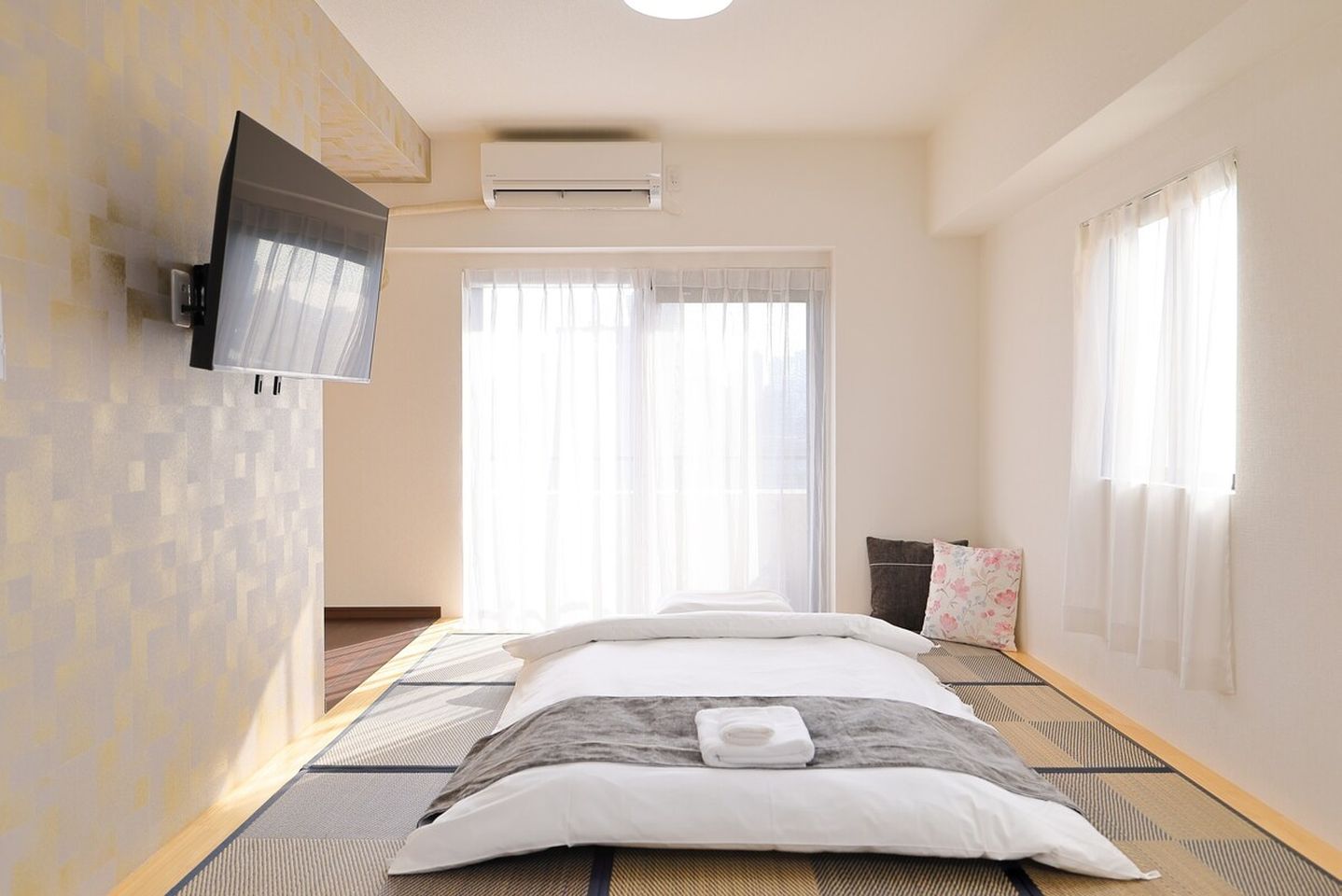 A futon bed on tatami mats in a Japanese-style room with a wall-mounted TV and bright windows.