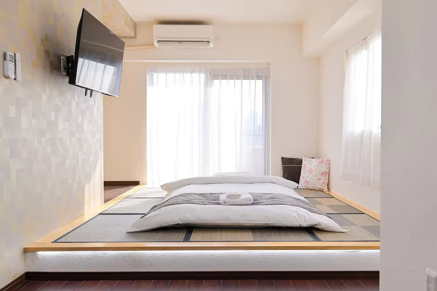 Futon bed on a raised tatami platform in a bright room with a wall-mounted TV and sheer curtains.